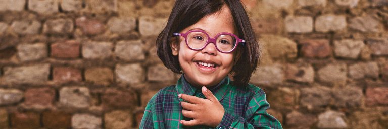 Girl with Down syndrome wearing glasses and standing in front of a brick wall