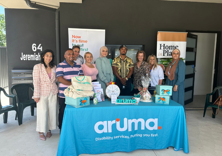 Aruma staff celebrating the completion of the new SDA homes in Werrington. They are standing behind an Aruma table with merchandise and signage.